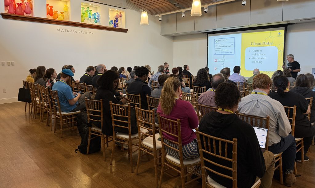 A full room of attendees seated in the Silverman Pavilion listens to presenter Andy, who stands beside a projected slide about using Salesforce Flow for clean data, featuring custom validations and automated cleaning.